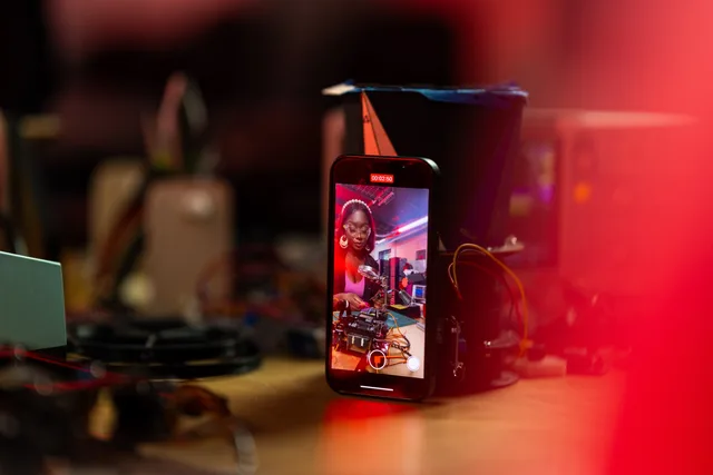 A woman records a tech tutorial using her smartphone on a desk filled with electronics. Video hosting and website delivery: embeds and playback